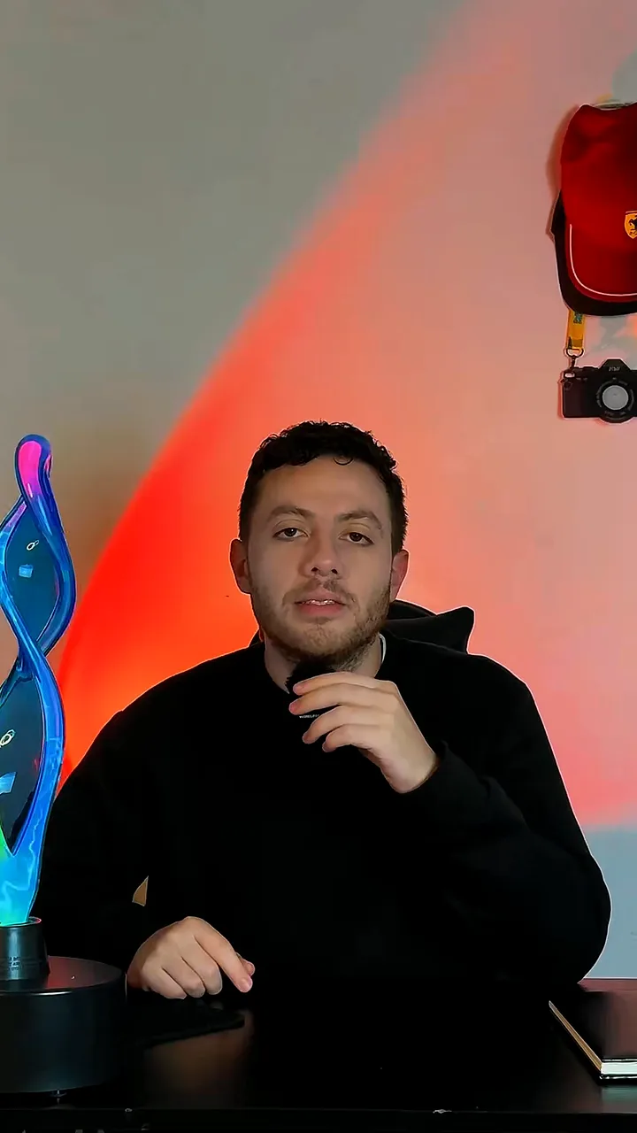 Man in a black sweater at a desk with his hand at his chin, a red glow lamp lighting the wall behind him