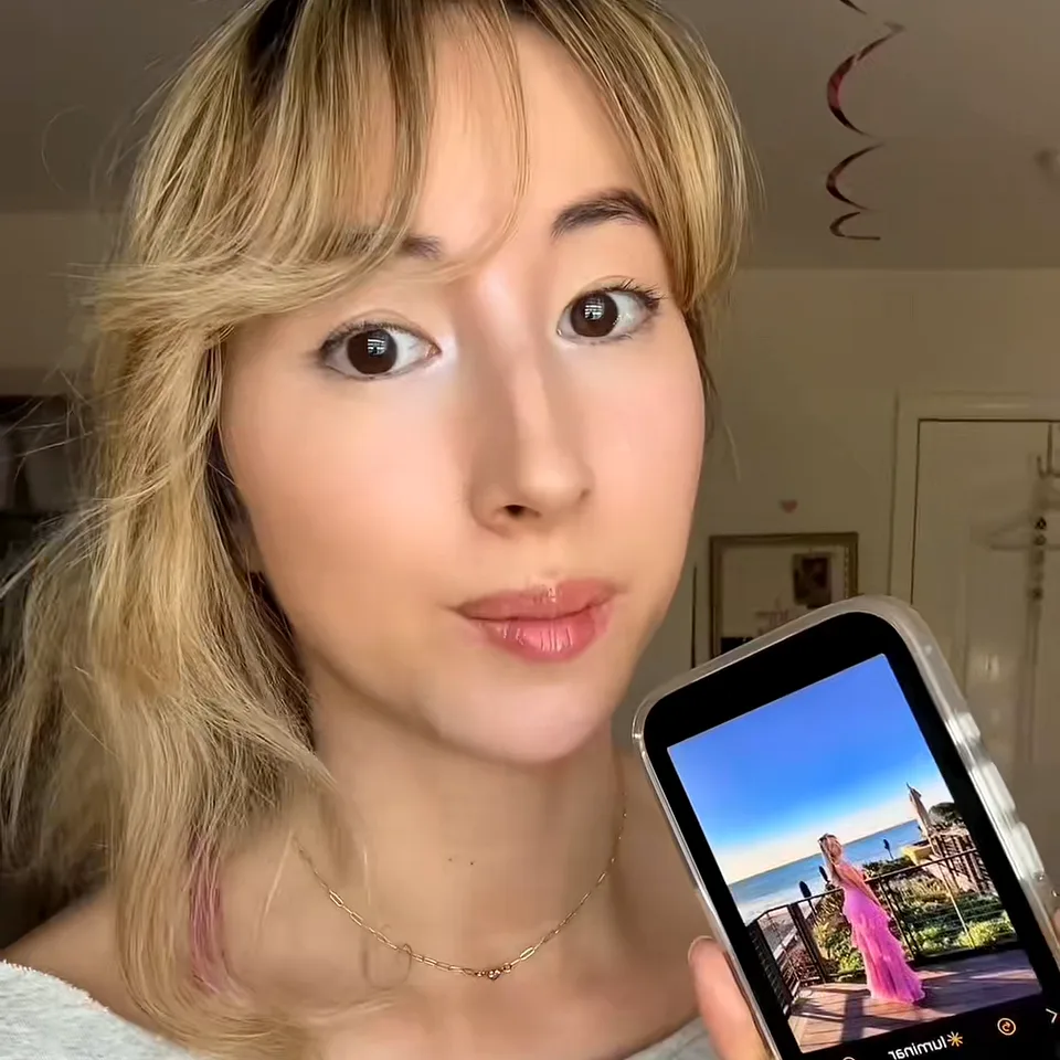 Creator with blonde bangs holding up her phone in the Luminar app with an edited seaside photo on screen