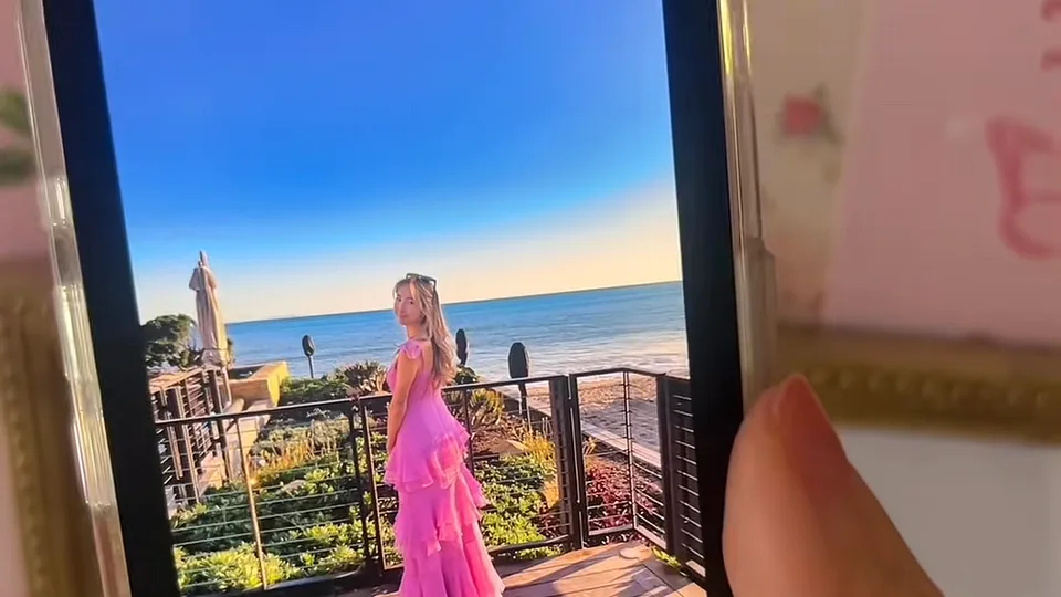 Phone screen showing a woman in a pink ruffled gown on a terrace above the sea at sunset