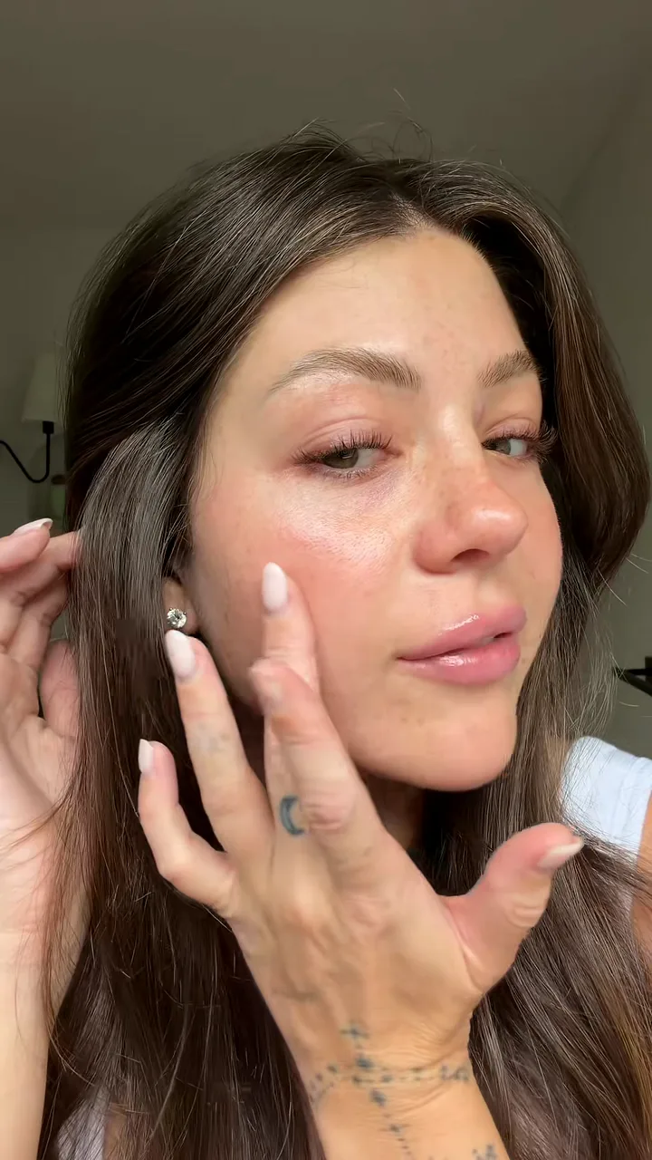 Freckled creator with long brown hair pressing her fingertips to her cheek to show dewy, glowing skin