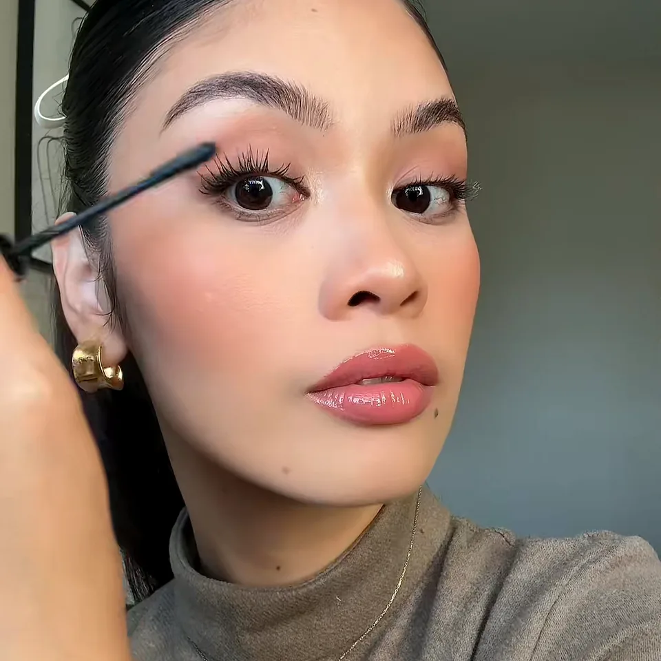 Creator with a sleek ponytail brushing mascara onto her upper lashes, gold earring in view