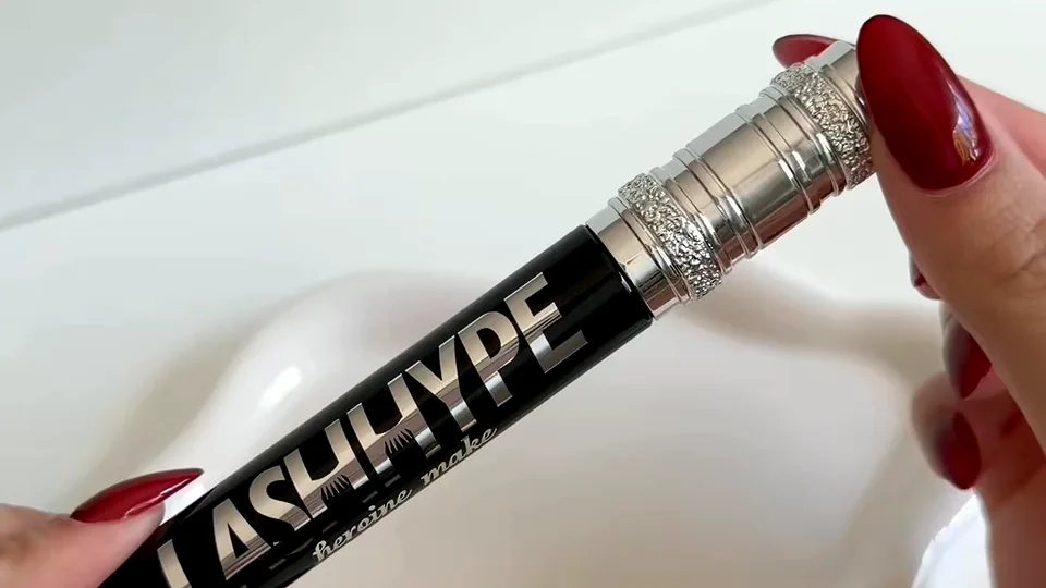 Hands with red nails holding a black and silver Lash Hype mascara tube over a white sink