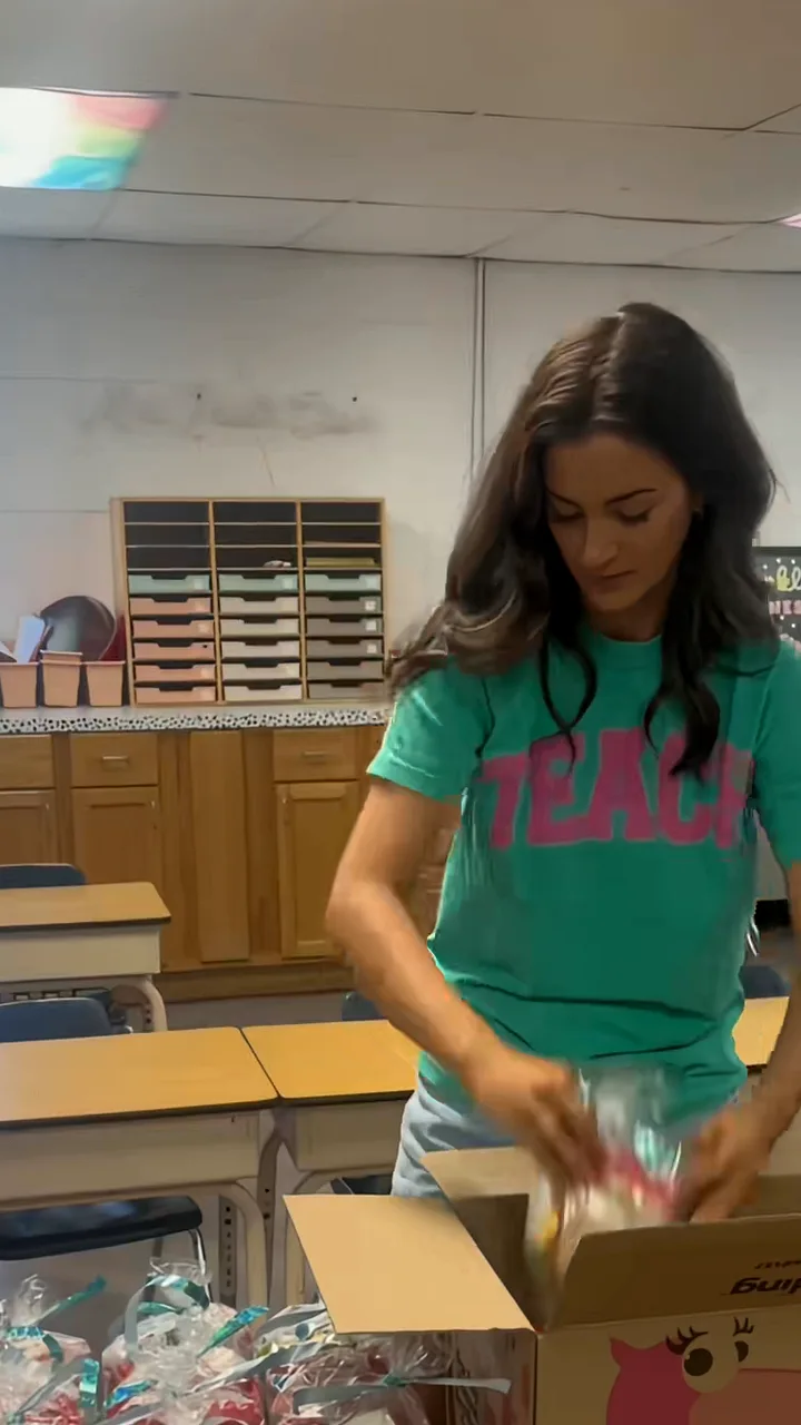Teacher in a green TEACH T-shirt unpacking wrapped party favours from a cardboard box in a classroom