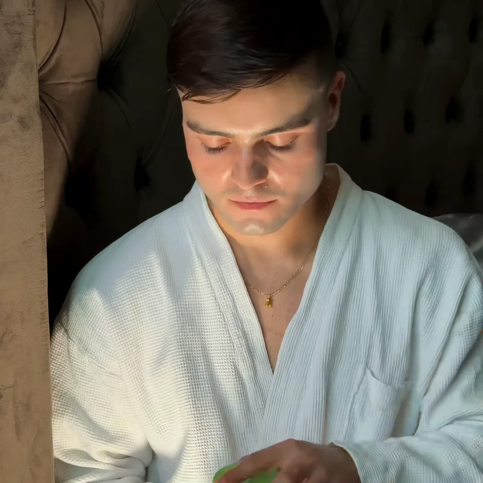 Man in a white waffle robe sitting in warm window light, eyes lowered to a green item in his hands