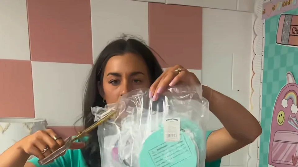 Woman in a teal top cutting open a plastic bag of craft kits with gold scissors in a decorated classroom