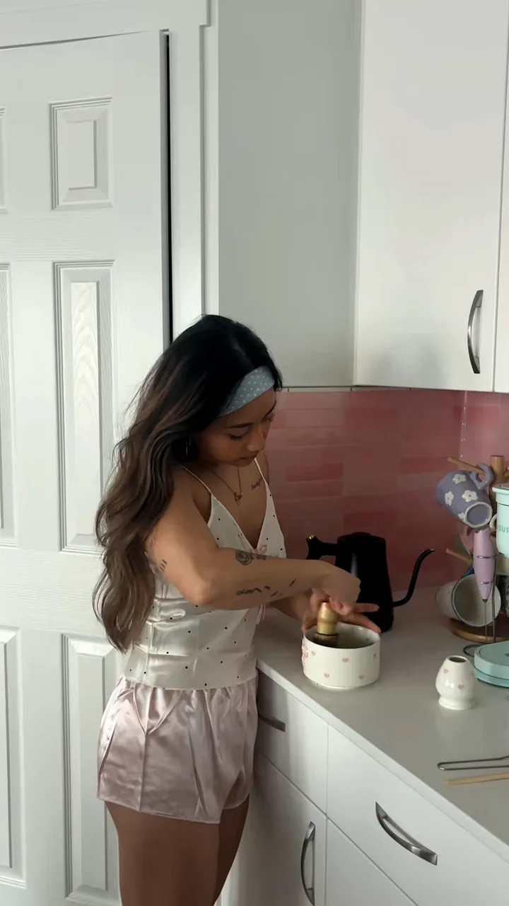 Woman in a satin cami and pink pyjama shorts whisking matcha in a bowl on a white kitchen counter
