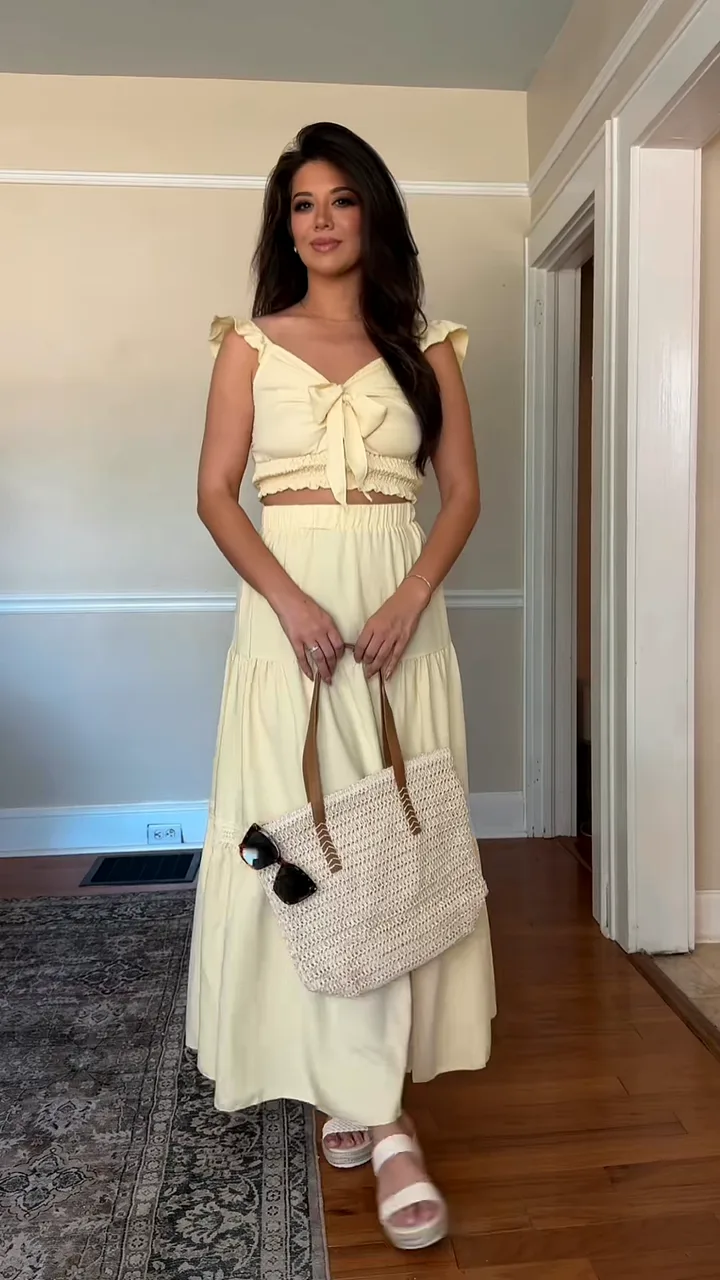 Woman in a pale yellow crop top and tiered maxi skirt holding a straw tote bag with sunglasses clipped on