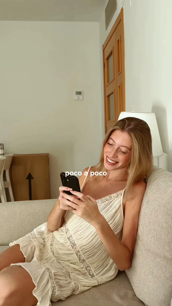 Woman in a cream sundress smiling at her phone while leaning back on a beige sofa at home