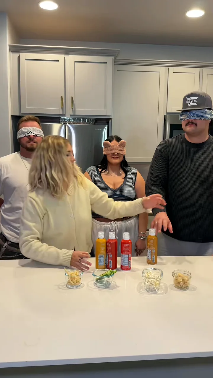 Woman guiding three blindfolded friends at a kitchen island set with popcorn spray bottles and snack cups