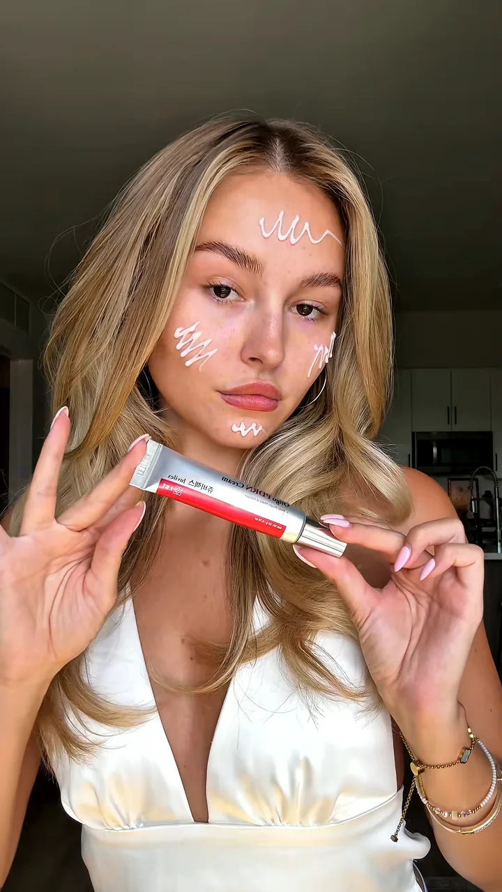 Creator with dots of white cream on her forehead and cheeks holding up a red and silver eye cream tube