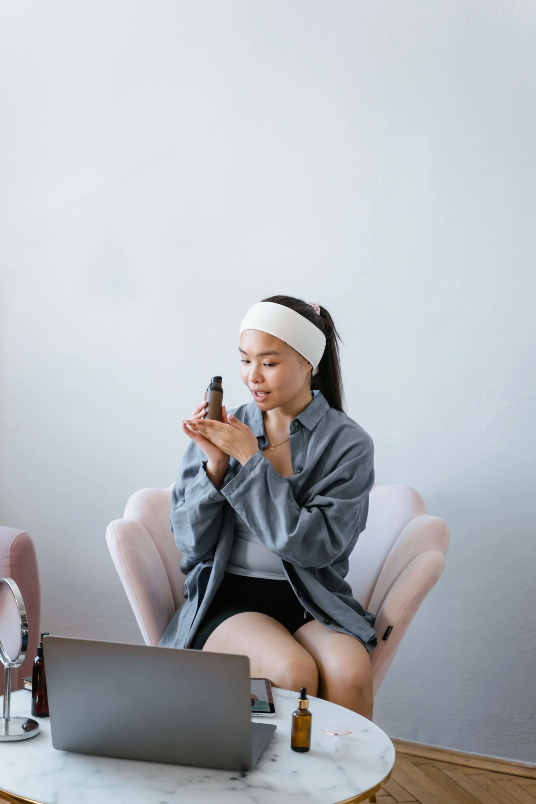 A woman in a white headband sits in a pink armchair and smells a small amber skincare bottle, with a laptop and a dropper bottle on the table in front of her.