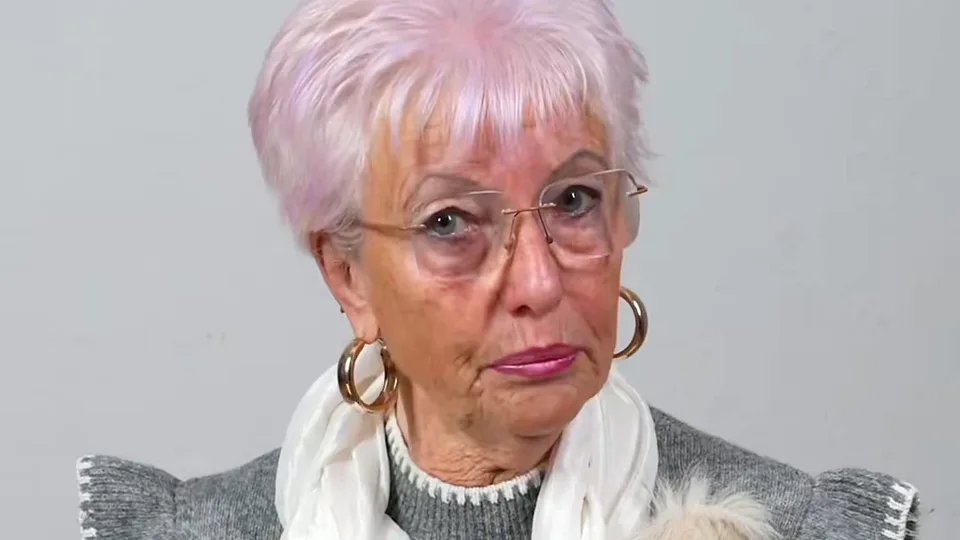 Woman with short lilac hair, glasses, gold hoop earrings and a white scarf listening against a grey wall