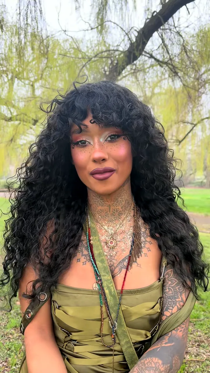 Woman with long black curls, face gems and layered necklaces smiling under a willow tree in a park