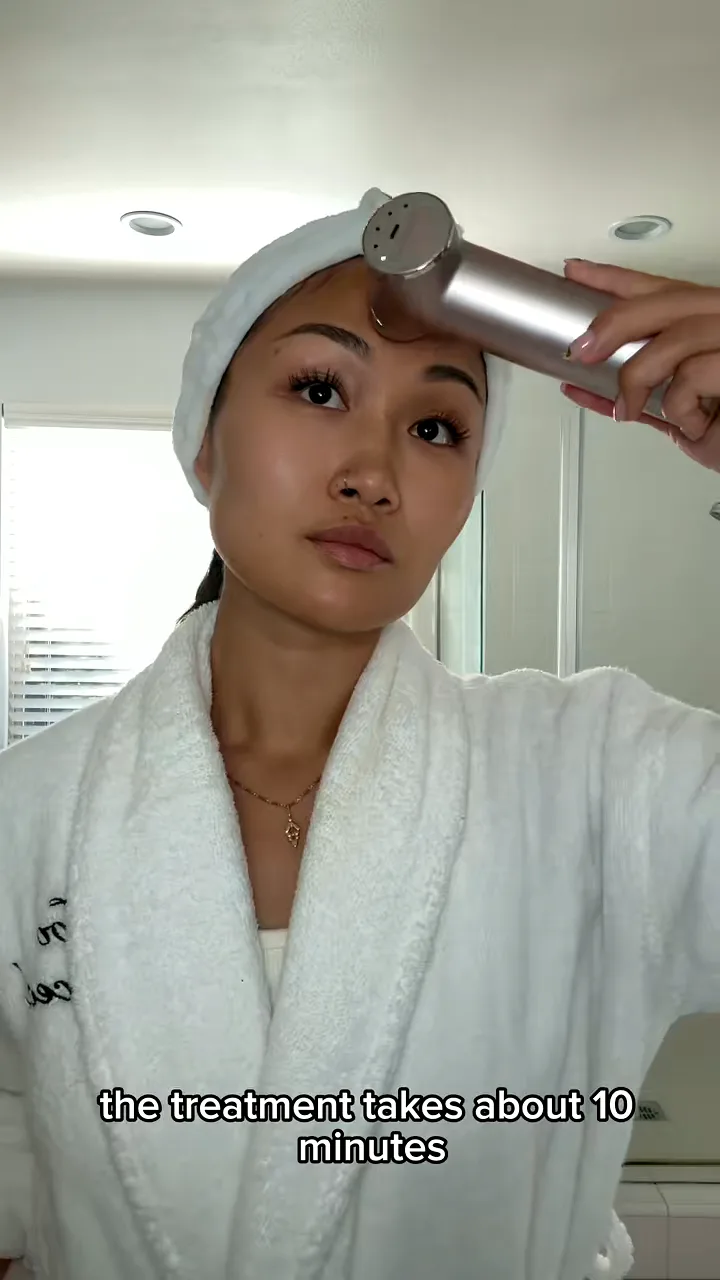 Woman in a white robe and towel headband holding a handheld skincare device to her forehead