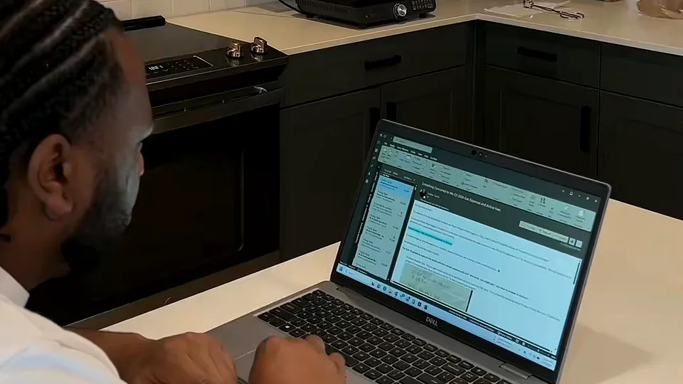 Man with braided hair typing on a Dell laptop at a white kitchen counter in a dark-cabinet kitchen